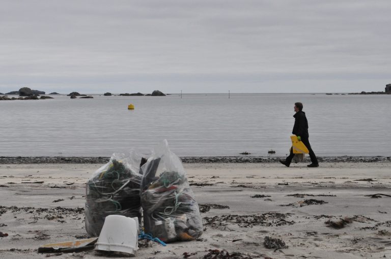 Sekker med marint avfall på stranda, rydder går i bakgrunnen